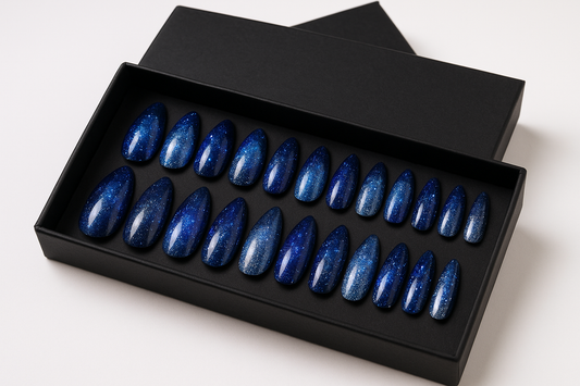 "Professional product photography of a set of 24 galaxy-themed press-on nails with deep space blue and silver stardust, holographic shimmer, glitter sparkles, elegant almond shapes, arranged in luxury black packaging box, clean white background, magical beauty product styling, realistic nail texture